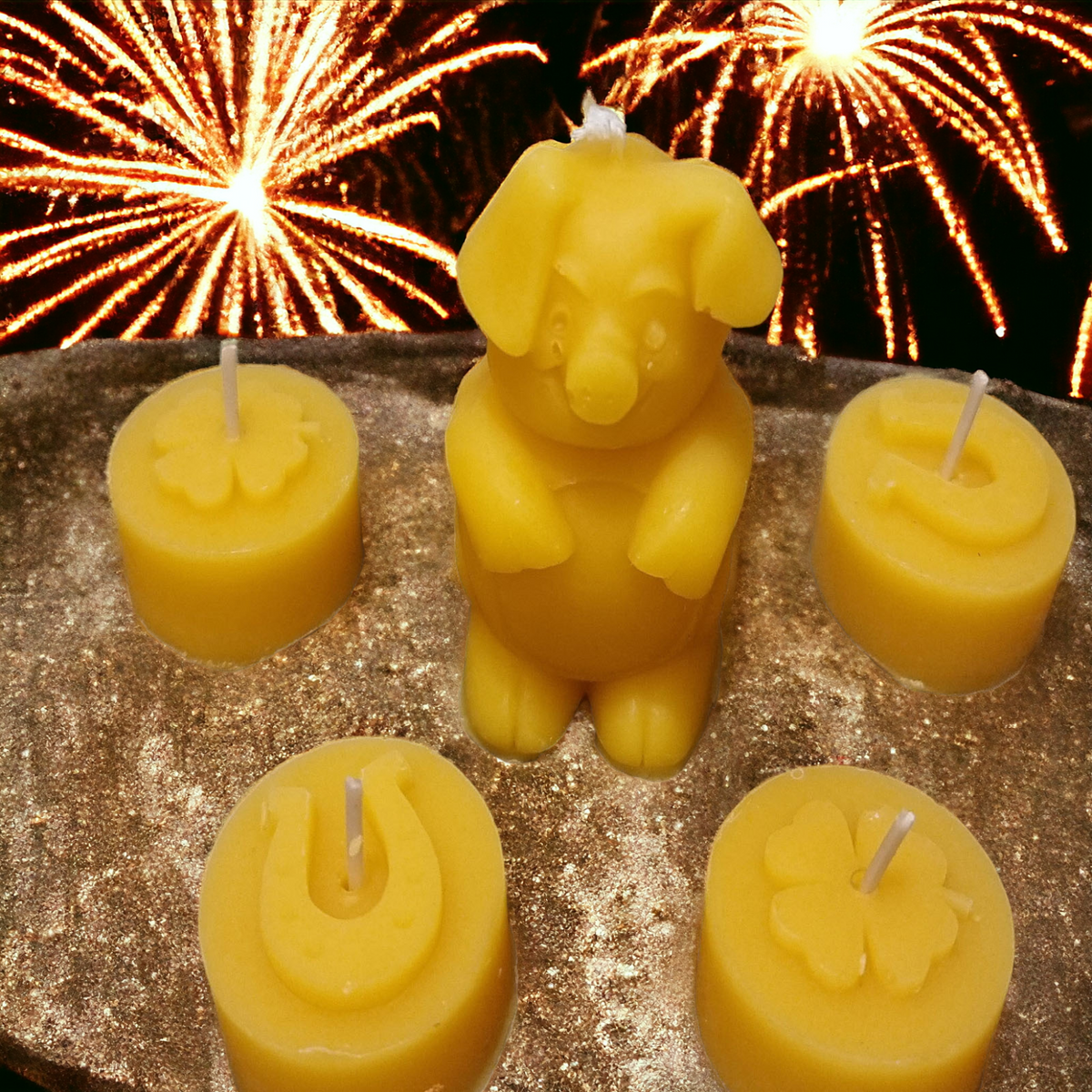 Ein Set aus fünf gelben Bienenwachskerzen mit Glückssymbolen ist auf einer golden glitzernden Unterfläche arrangiert. In der Mitte steht eine stehende Glücksschweinchen-Kerze, umgeben von kleinen Stumpenkerzen mit erhabenen Hufeisen- und Kleeblatt-Motiven. Im Hintergrund ist ein farbenprächtiges Silvester-Feuerwerk am Nachthimmel zu sehen.