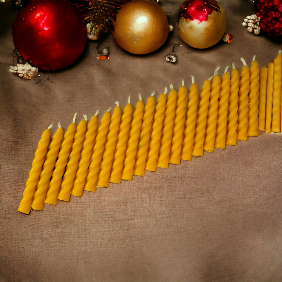 Eine Reihe von vielen handgefertigten, spiralförmig gedrehten Bienenwachskerzen in Gelb, die auf einem textilen Untergrund vor weihnachtlicher Dekoration mit roten und goldenen Christbaumkugeln liegen.