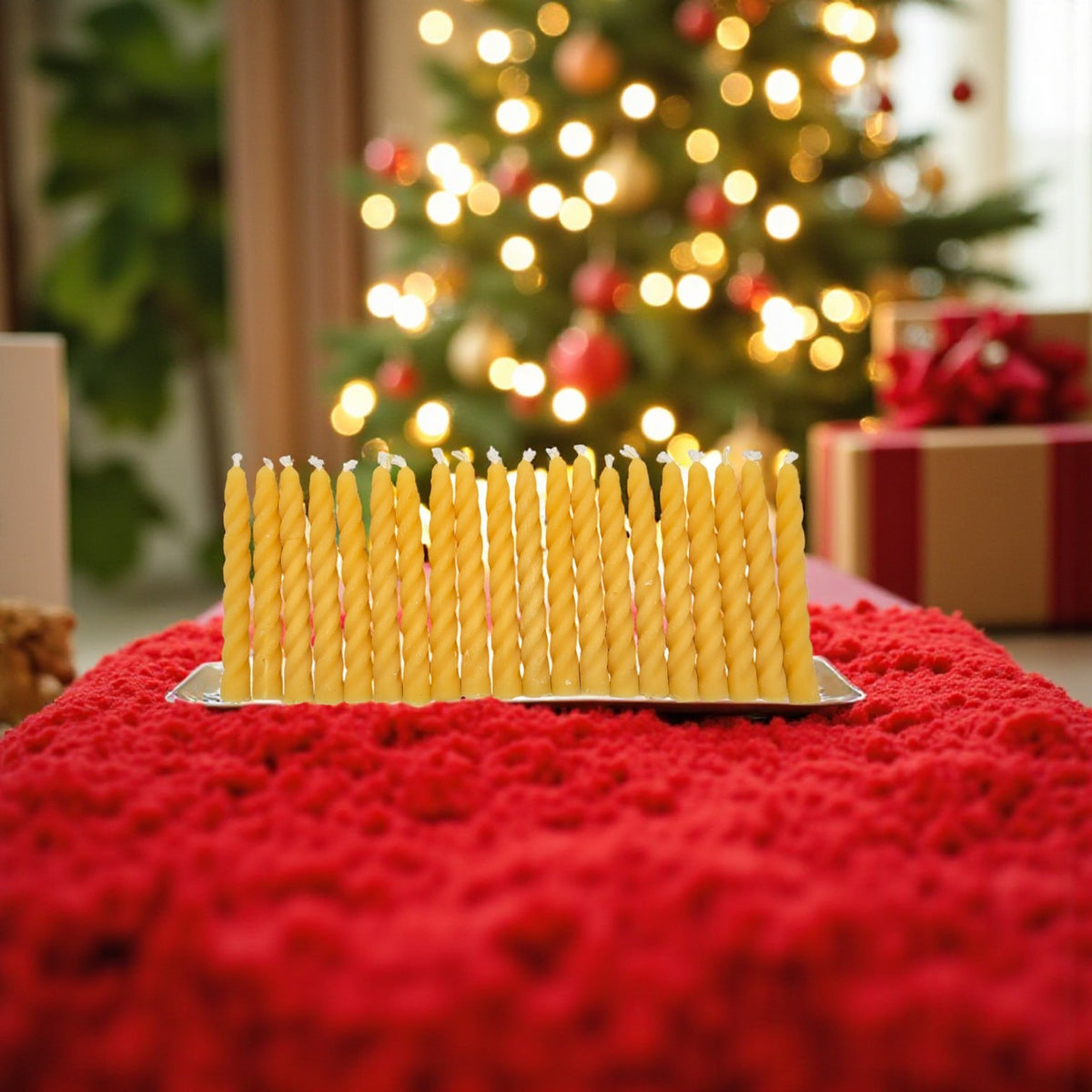 Ein Set aus handgefertigten, spiralförmig gedrehten Bienenwachs-Weihnachtsbaumkerzen, aufgereiht auf einer roten Decke vor einem festlich geschmückten Tannenbaum mit warmen Lichtern.