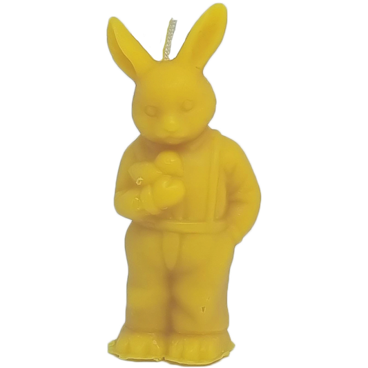 Eine detailreiche Kerze aus gelbem Bienenwachs in Form eines aufrecht stehenden Osterhasen vor weißem Hintergrund. Der Hase trägt eine Latzhose mit Hosenträgern, hält einen kleinen Gegenstand in den Pfoten, hat lange, aufgestellte Ohren und an der Oberseite ragt ein Docht heraus.