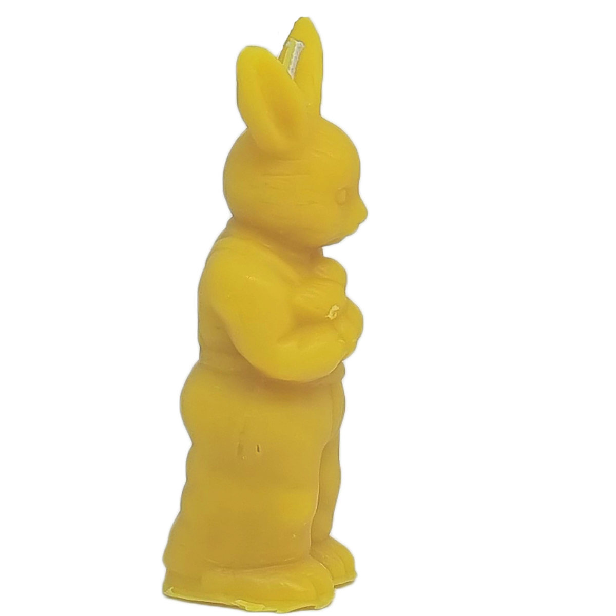 Eine detailreiche Kerze aus gelbem Bienenwachs in Form eines aufrecht stehenden Osterhasen in der Seitenansicht vor weißem Hintergrund. Der Hase trägt eine Latzhose, hält einen kleinen Gegenstand in den Pfoten und hat lange, aufgestellte Ohren, an deren Zwischenraum der Docht zu erkennen ist.