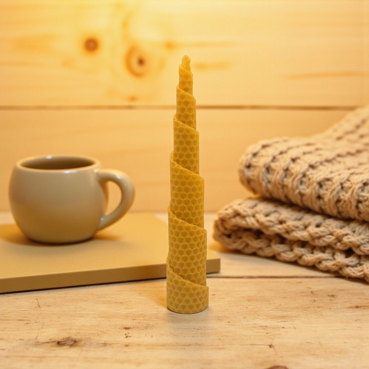 Eine spiralförmig gedrehte Bienenwachskerze mit Wabenstruktur steht aufrecht auf einer hellen Holzoberfläche. Im gemütlichen Hintergrund sind eine Keramiktasse auf einem Buch und ein gefalteter, heller Strickpullover zu sehen.