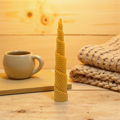 Eine spiralförmig gedrehte Bienenwachskerze mit Wabenstruktur steht aufrecht auf einer hellen Holzoberfläche. Im gemütlichen Hintergrund sind eine Keramiktasse auf einem Buch und ein gefalteter, heller Strickpullover zu sehen.