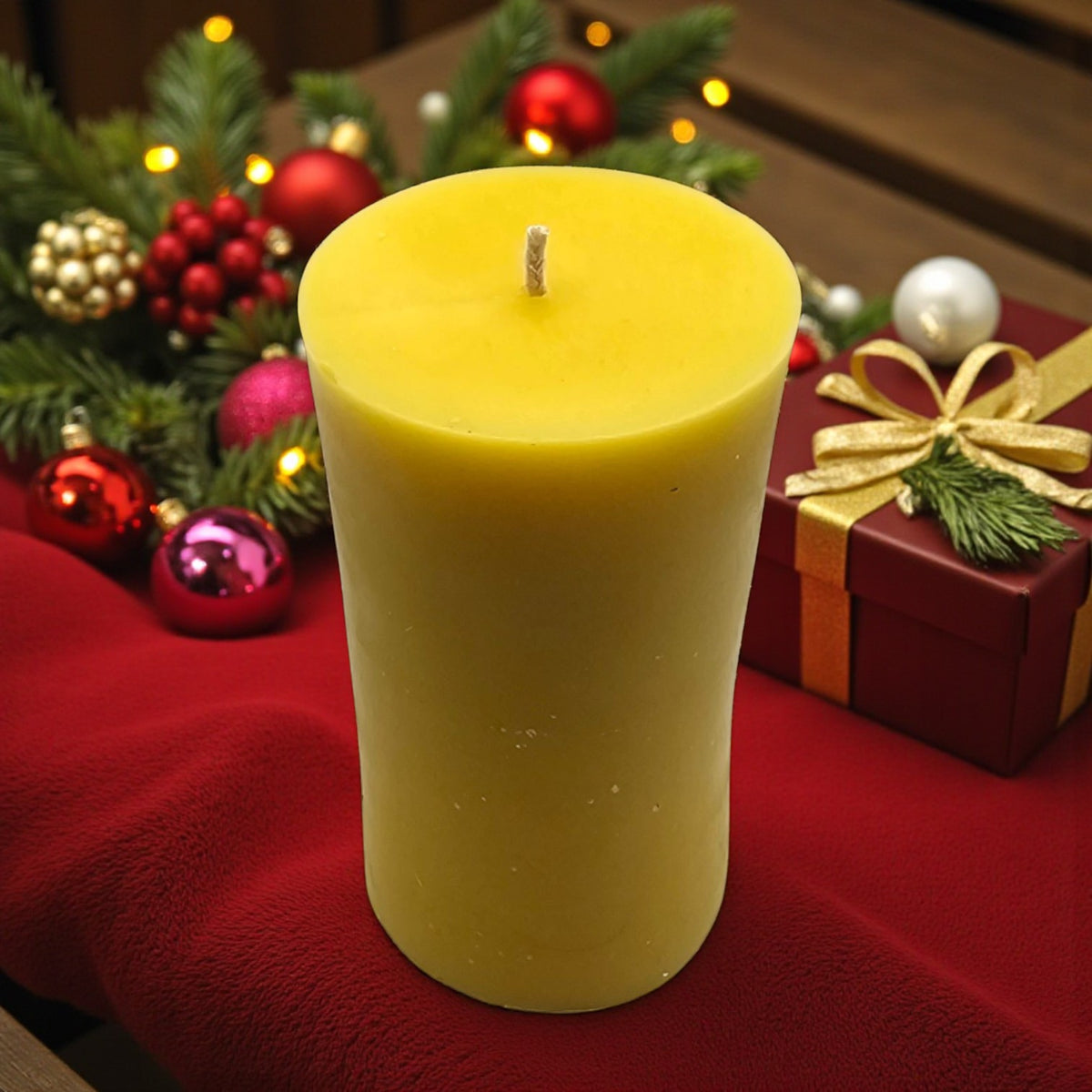 Eine glatte Stumpenkerze aus gelbem Bienenwachs auf rotem Stoff, festlich dekoriert mit roten und goldenen Weihnachtskugeln, Tannenzweigen und einem kleinen Geschenk im Hintergrund.
