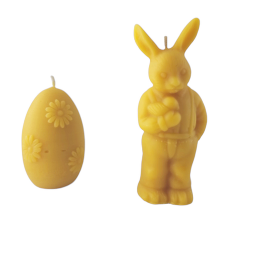 Zwei gelbe Bienenwachskerzen mit österlichen Motiven stehen nebeneinander vor einem weißen Hintergrund. Links ist eine eiförmige Kerze mit erhabenen Blümchen-Mustern zu sehen, rechts eine detailreiche Kerze in Form eines stehenden Osterhasen in Latzhose. Auf der Oberseite beider Kerzen befindet sich jeweils ein kleiner Docht.