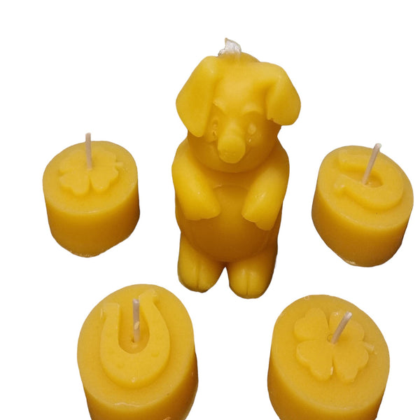 Ein Set aus fünf kleinen Kerzen aus gelbem Bienenwachs mit typischen Glückssymbolen vor weißem Hintergrund. In der Mitte steht eine stehende Glücksschweinchen-Kerze, umgeben von vier runden Teelichtern beziehungsweise kleinen Stumpenkerzen, die mit erhabenen Motiven von Hufeisen und vierblättrigen Kleeblättern verziert sind. Auf jeder Kerzenoberseite befindet sich ein kleiner Docht.